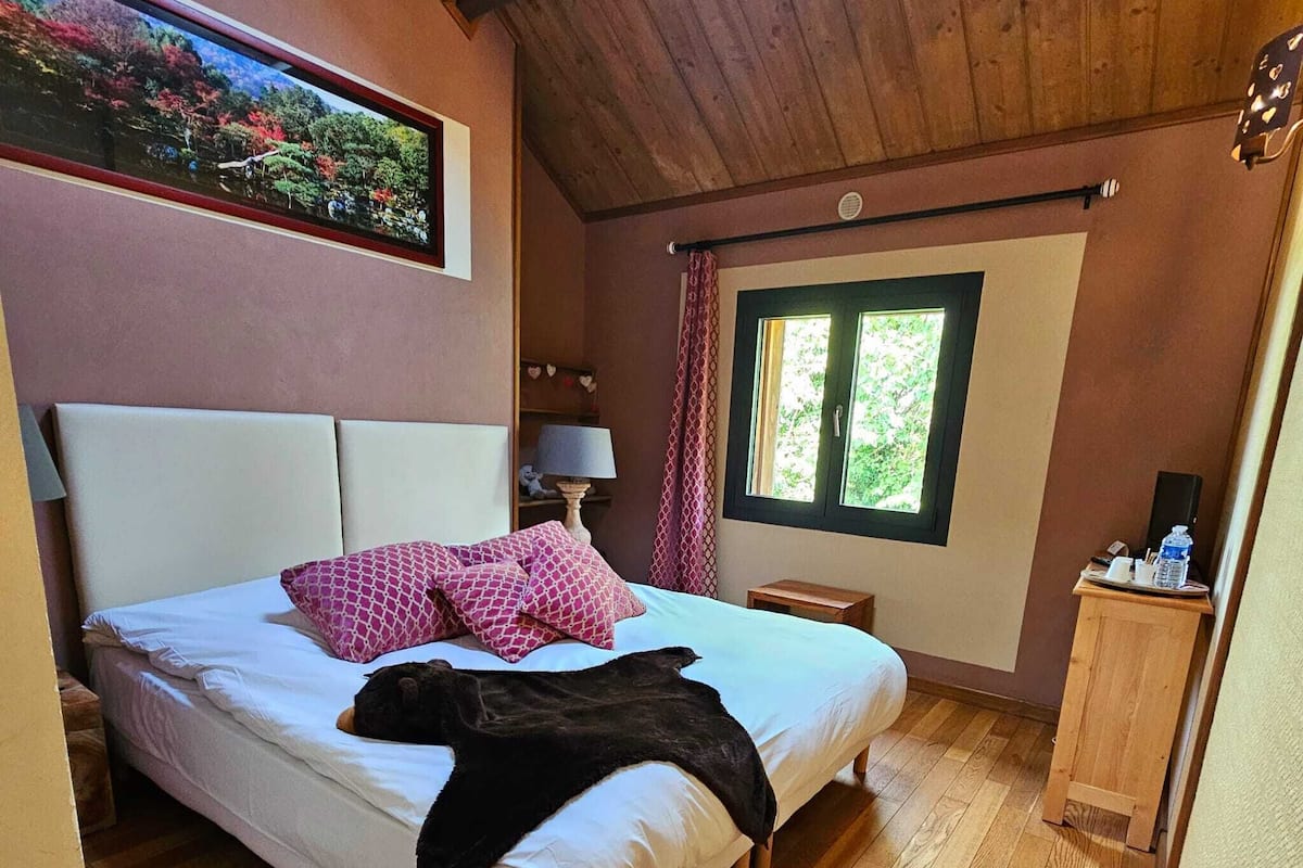 Een slaapkamer met een bed, nachtkastje, lamp, raam en een houten plafond.