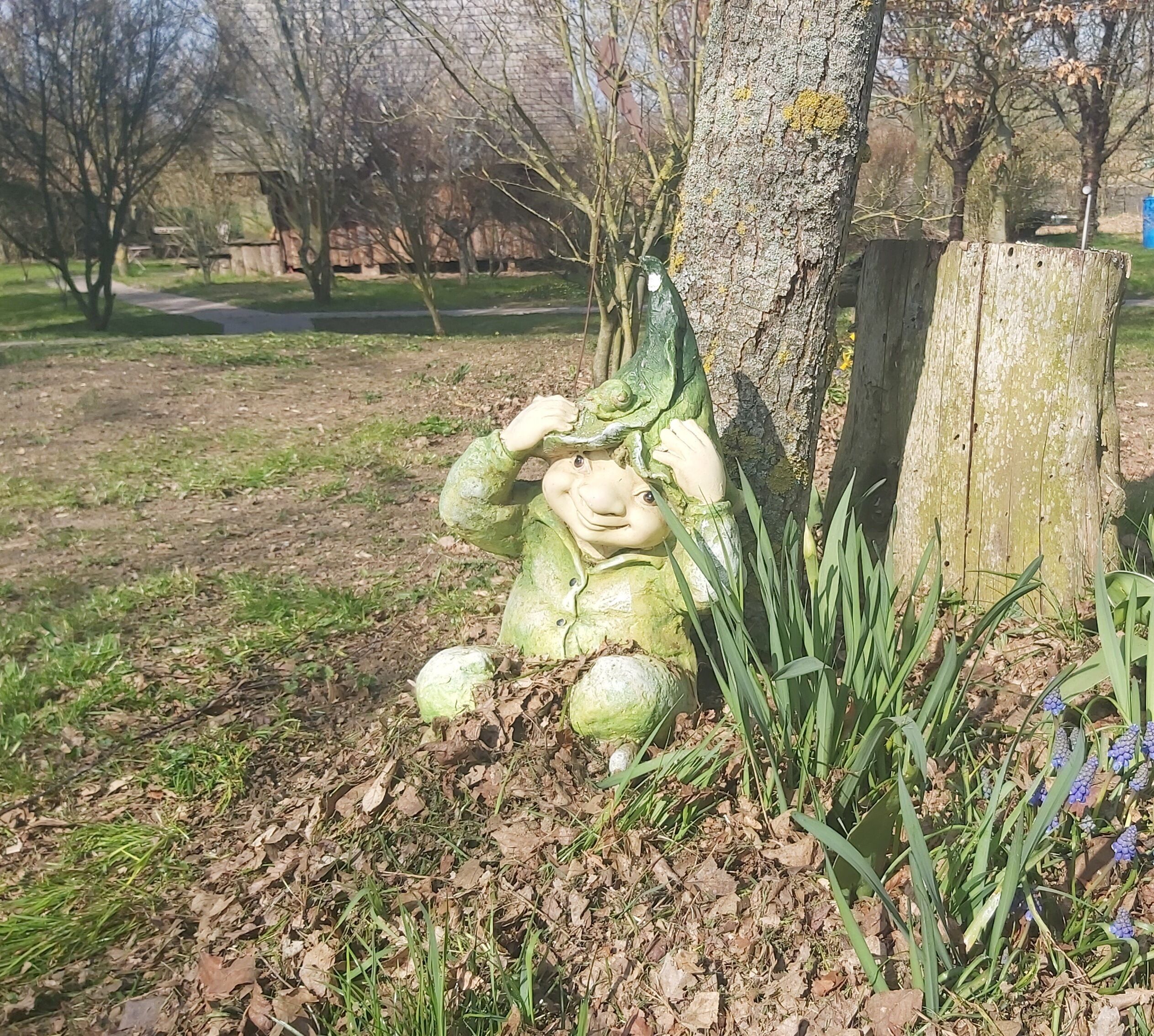A garden statue of a figure in a green costume sitting by a tree.