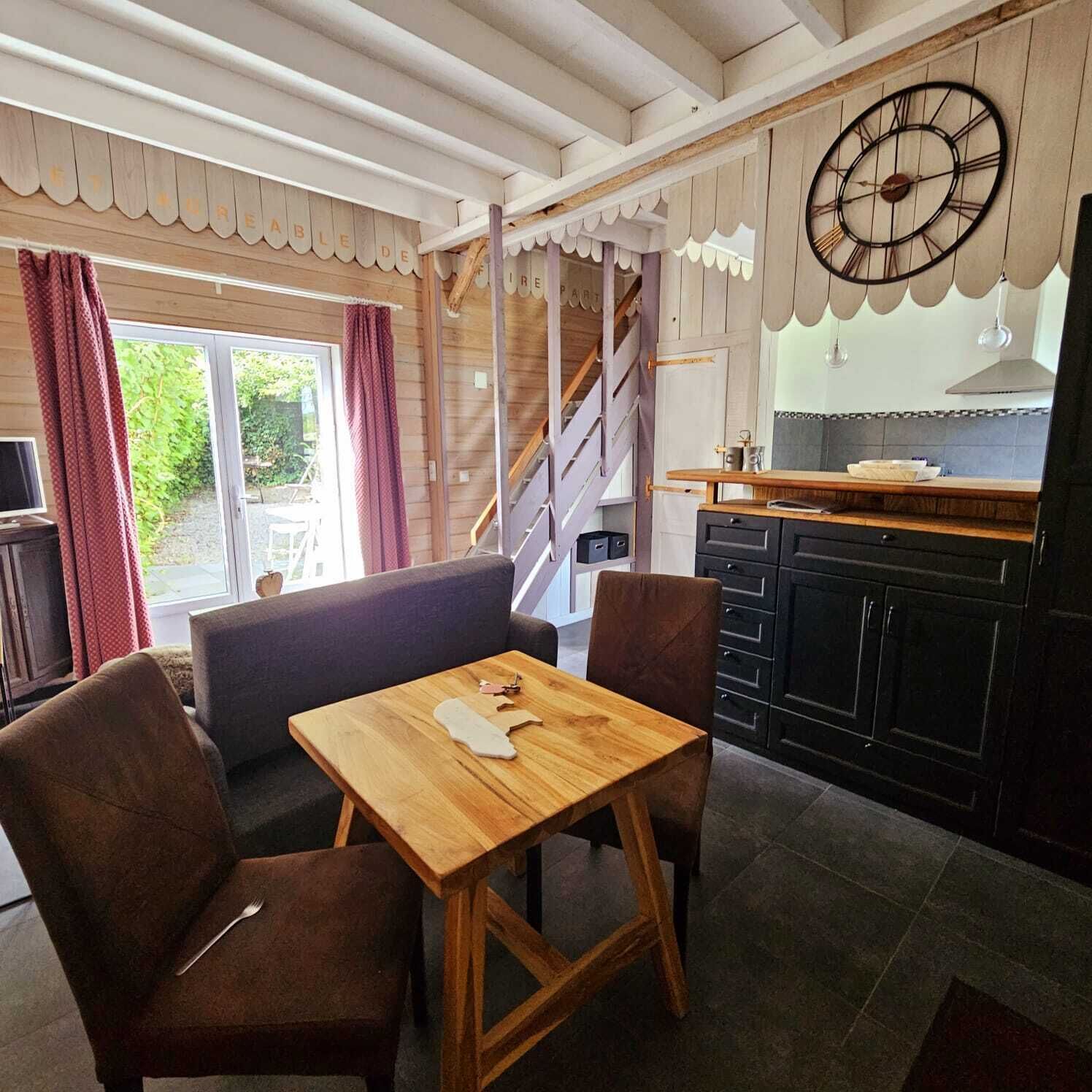 Een gezellige kamer met een houten tafel en stoelen, een grote klok aan de muur, en een keukenhoek met kastjes en een raam.