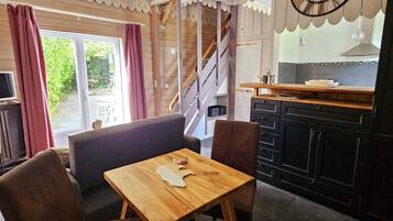 Een gezellige kamer met een houten tafel en stoelen, een grote klok aan de muur, en een keukenhoek met kastjes en een raam.