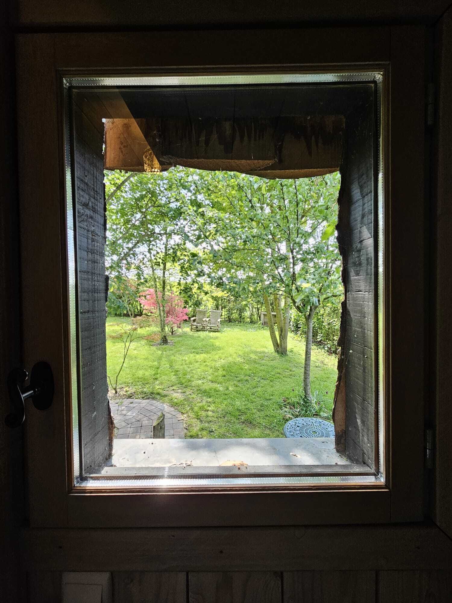 Una ventana con vista a un jardín, que incluye césped, árboles y un banco de madera.