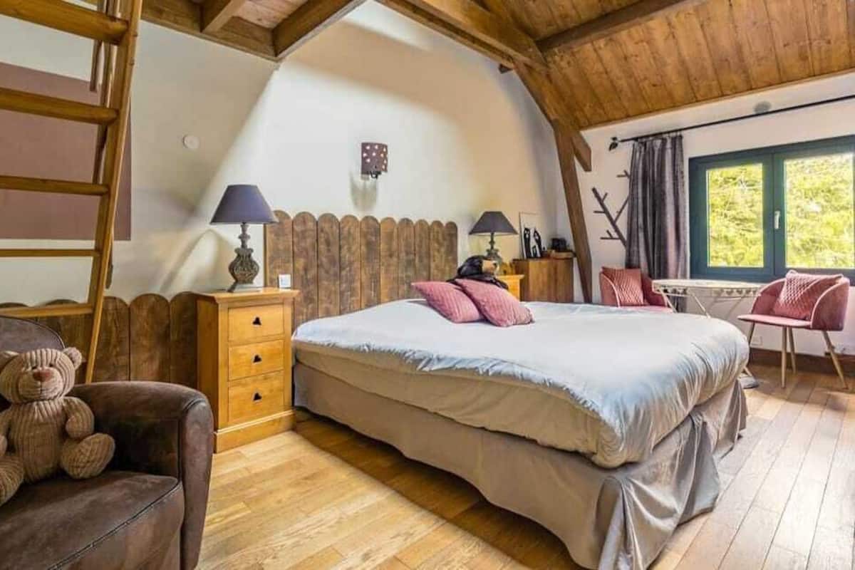 Een knus slaapkamer met een houten plafond, een bed met kussens, een stoel met een teddybeer, en een raam met gordijnen.