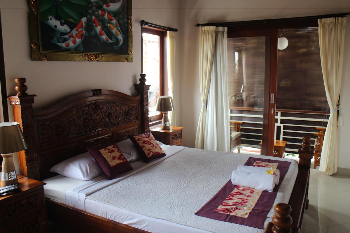 A neatly made bed with a white comforter and a purple and red floral throw. There is a wooden headboard, a nightstand with a lamp, and a painting of koi fish on the wall.