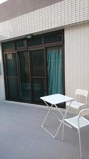 Un edificio con fachada de azulejos, puerta de vidrio corredera con cortinas y dos sillas blancas de metal en un patio con suelo de baldosas.