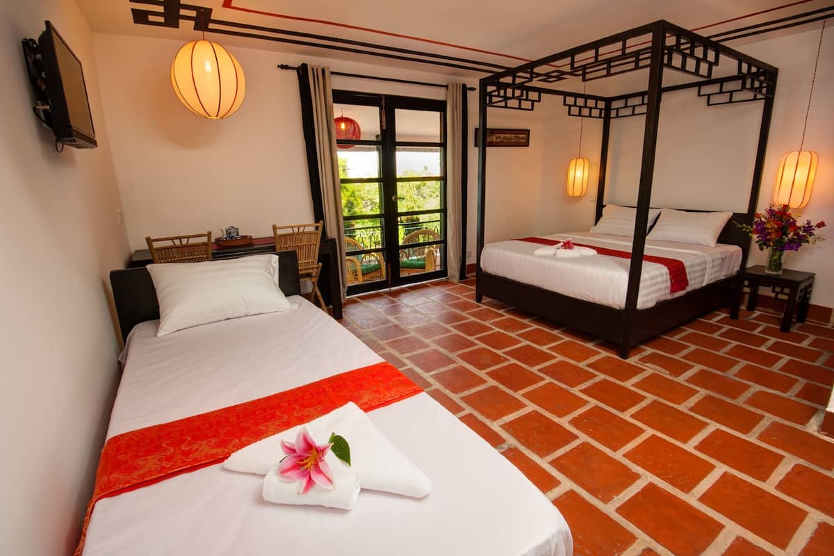 Une chambre d’hôtel dotée d’un grand lit, d’un lit plus petit, d’un balcon avec des meubles en osier et d’un lit à baldaquin de style asiatique traditionnel.