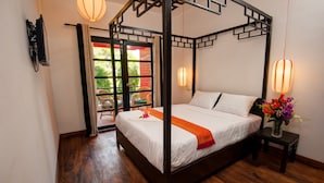 A four-poster bed with a white bedspread and orange throw, a small bedside table with a vase of flowers, and a view of the outdoors through a sliding door. - Villa Silk Road Hotel (Phnom Penh)