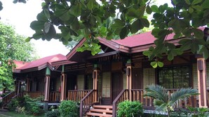 Une maison traditionnelle en bois, avec un toit rouge, une véranda dotée d’un escalier et un jardin bien entretenu où poussent différentes espèces de plantes.