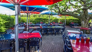 An outdoor dining area with tables and chairs under red and blue umbrellas, surrounded by greenery. - Stars and Stripes - Motel Steighof (Brutten)