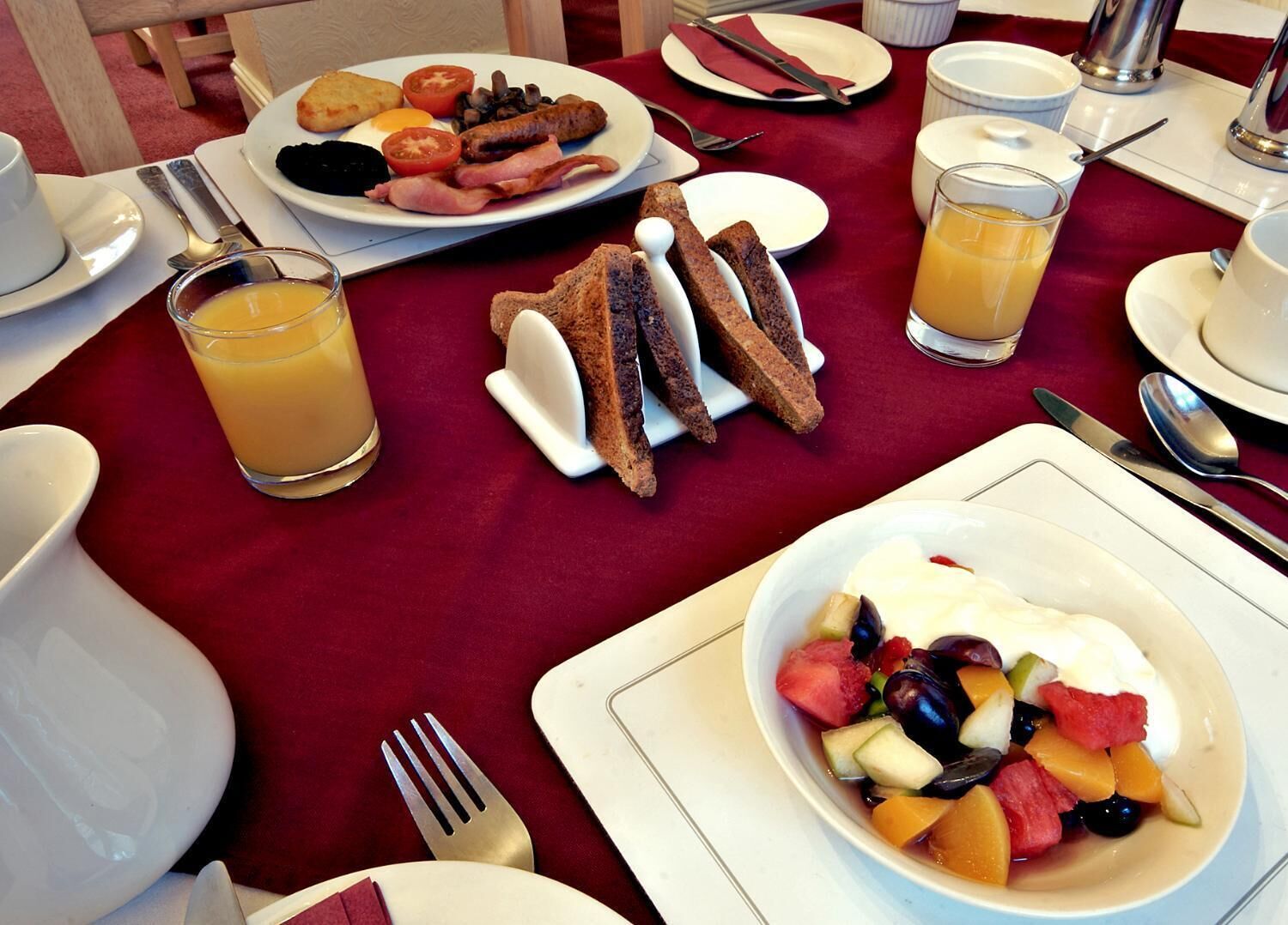 A breakfast setting with plates of eggs, sausages, tomatoes, and toast, along with a bowl of fruit salad and orange juice.