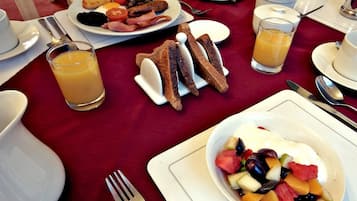 A breakfast setting with plates of eggs, sausages, tomatoes, and toast, along with a bowl of fruit salad and orange juice.