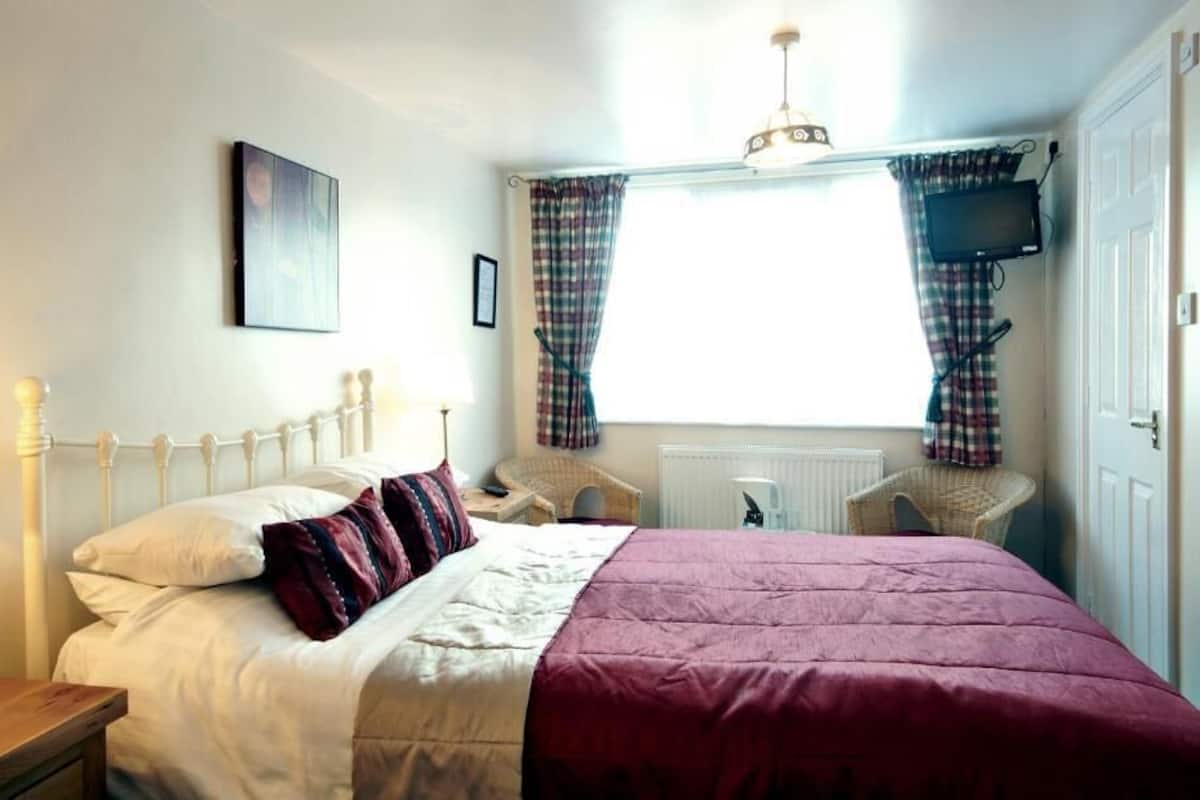 A bedroom with a bed, a bedside table with a lamp, a window with curtains, a television on the wall, and a radiator.