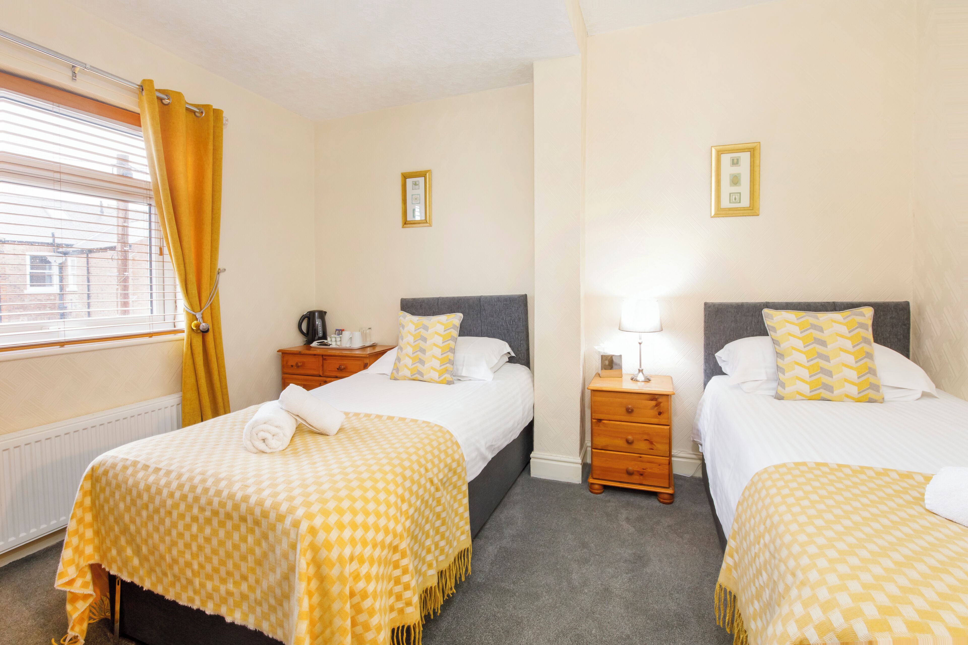 Habitación de hotel con dos camas, cada una con cubrecama y almohadas a cuadros, una mesita de noche con lámpara y una ventana con cortinas.