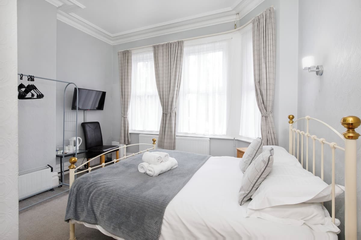 A neatly made bed with white and gray bedding, a television on a stand, a chair, and a radiator.