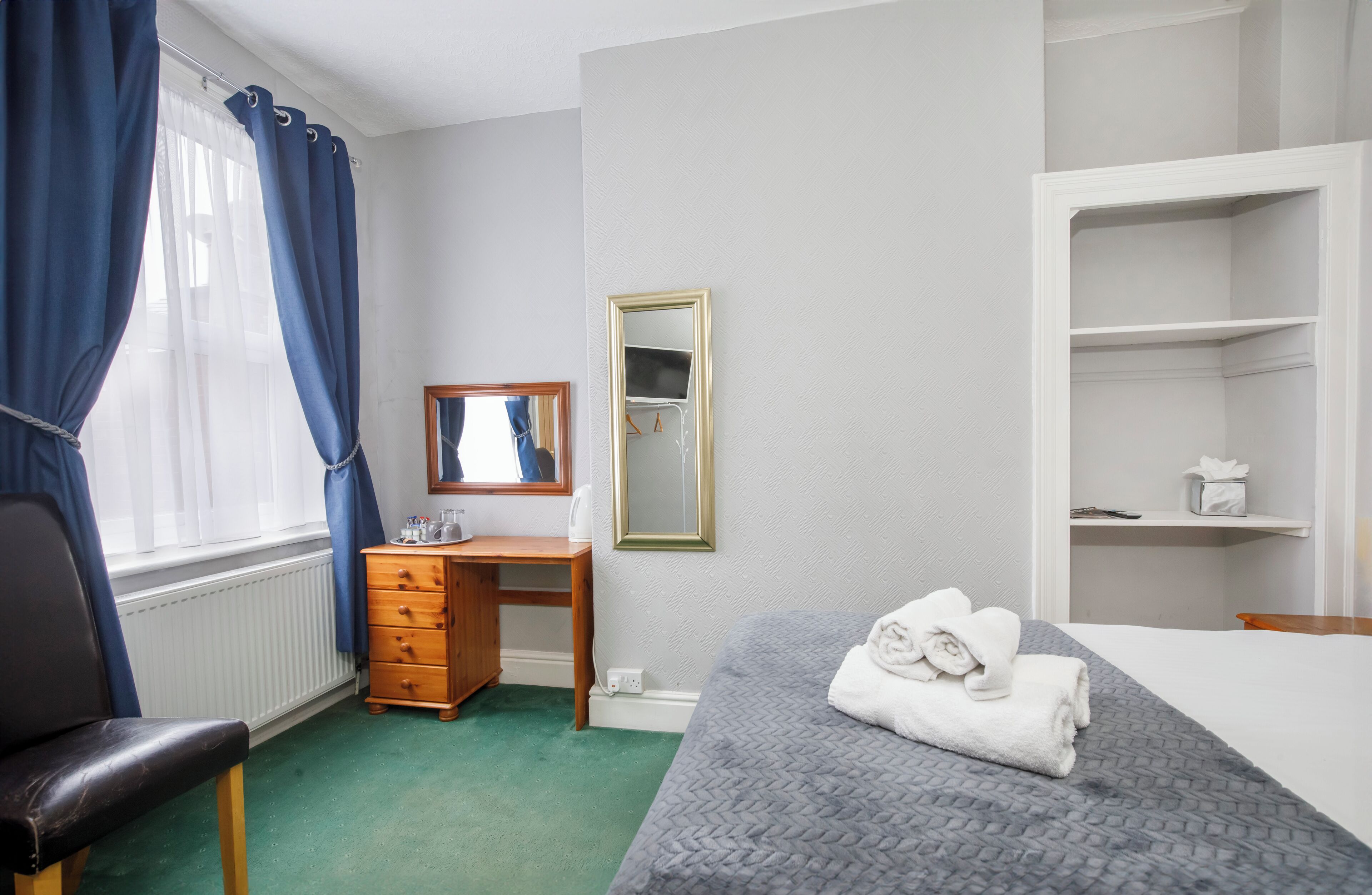 A bedroom with a bed, a wooden dresser, a desk, a mirror, and a radiator.