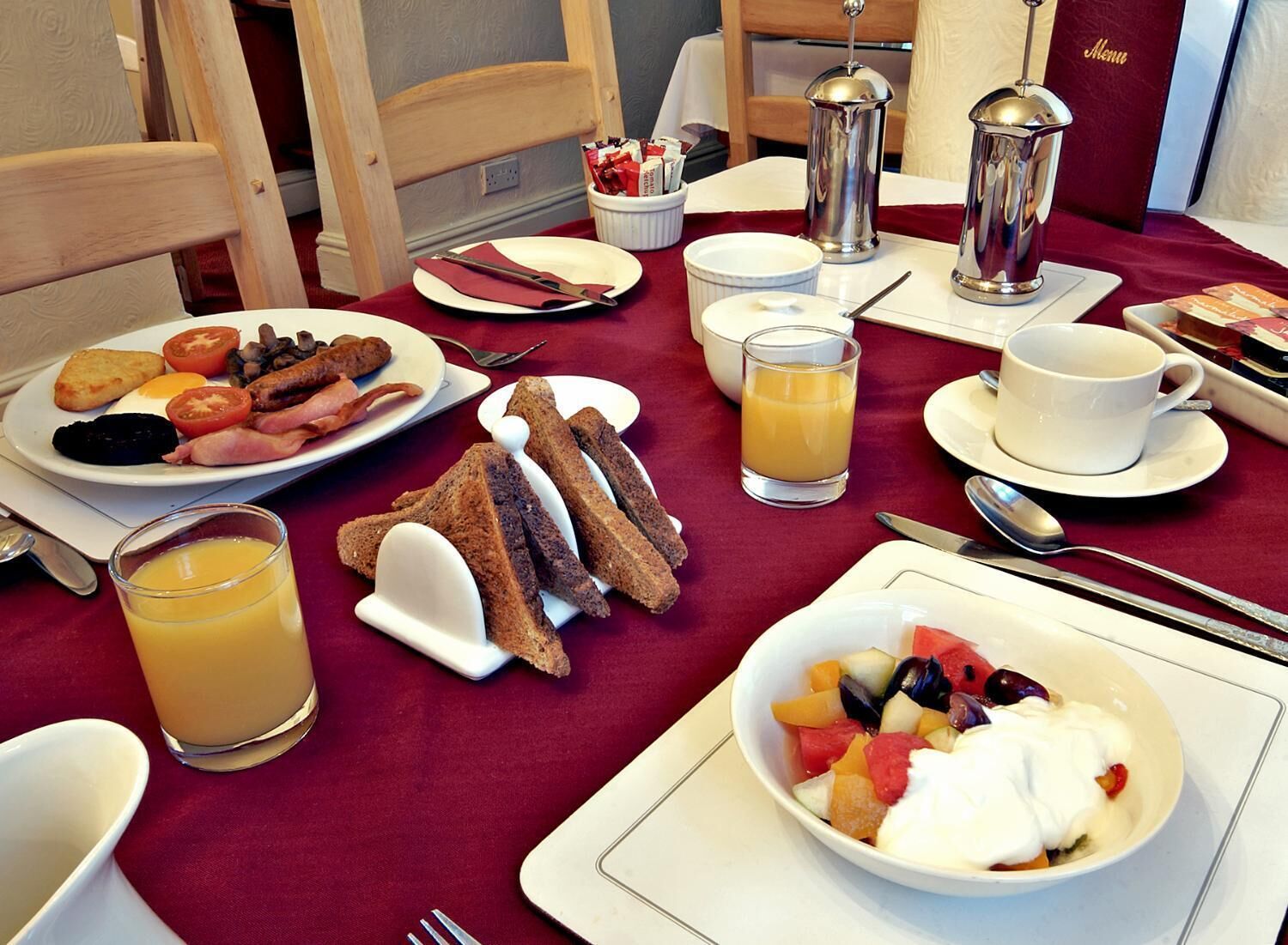 A breakfast setting with plates of eggs, bacon, toast, and fruit salad, along with juice and coffee.