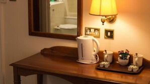 A hotel room with a wooden table, a white electric kettle, a tray with cups and a bowl of tea bags, and a wall-mounted lamp. - Abbey Lodge Hotel (High Wycombe)