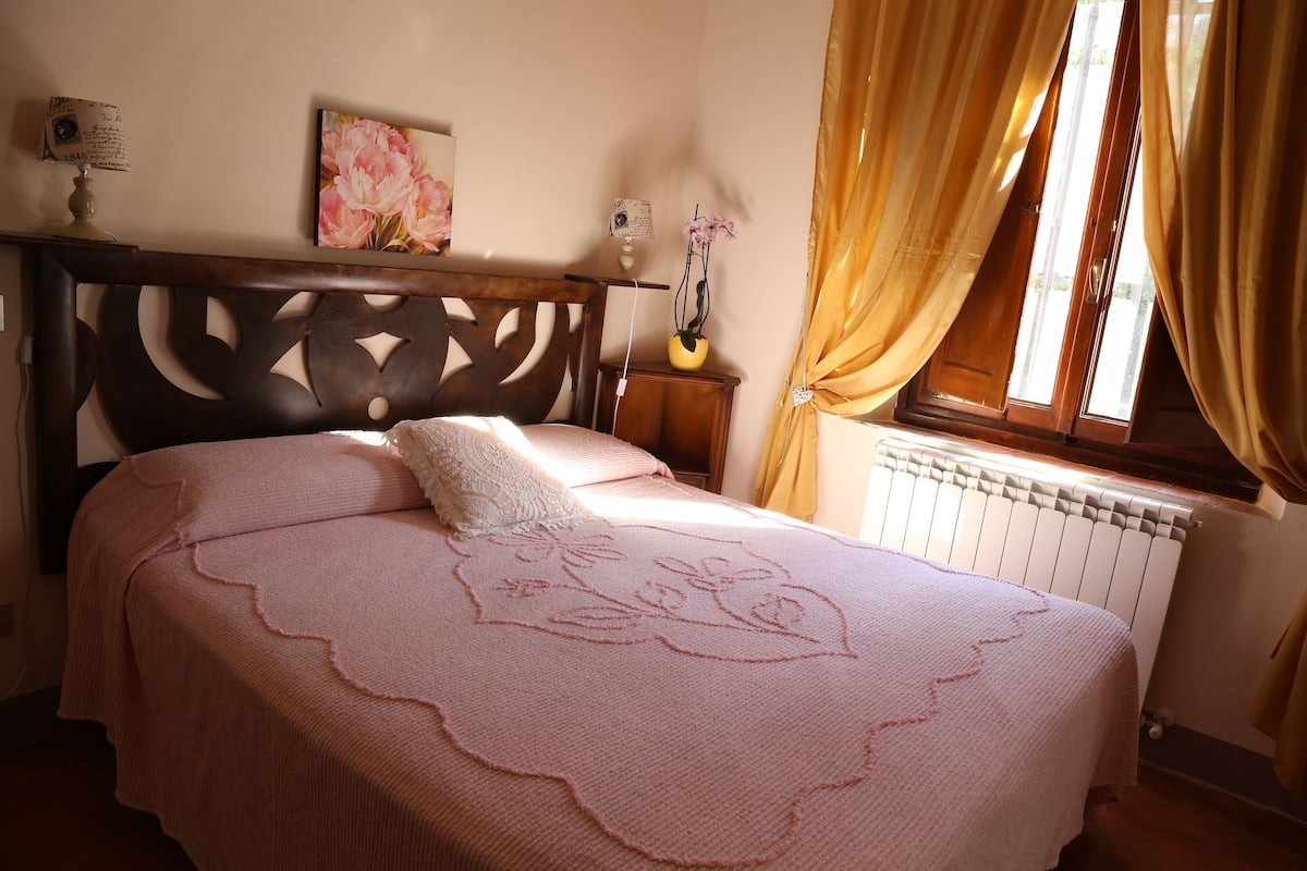 Un dormitorio con cabecera de madera, colcha rosa con bordados florales, una mesita de noche con un florero y una ventana con cortinas.