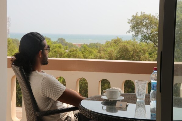 Una persona sentada a una mesa redonda con una taza y un vaso, contemplando una vista panorámica de árboles y el mar.