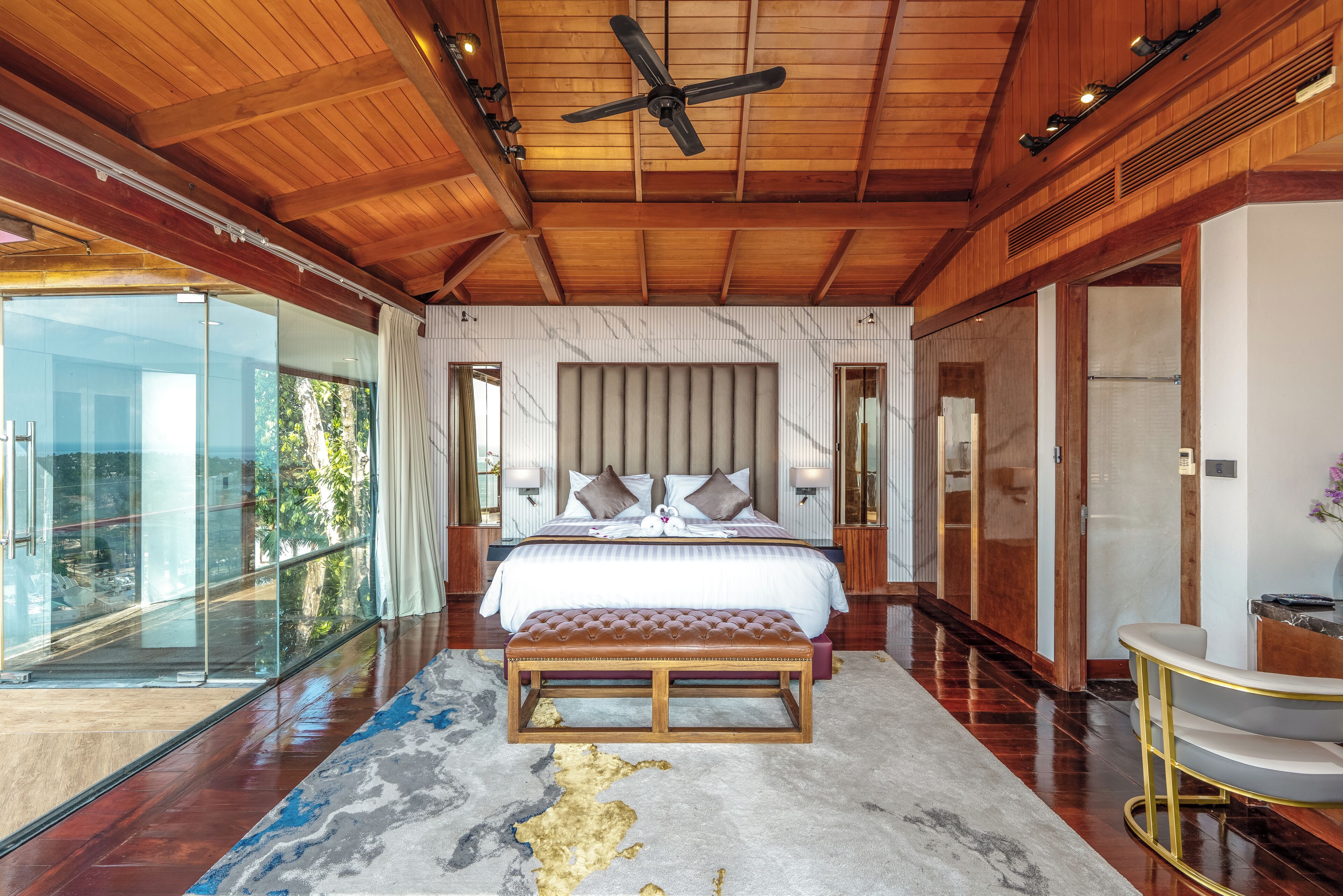A bedroom with a large bed, a bench, a ceiling fan, and a view through a glass door.