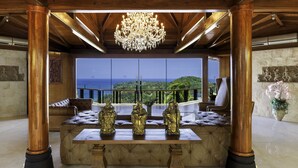 Een luxueuze kamer met een groot raam dat uitzicht geeft op zee, een kroonluchter en een zithoek met een tafel waarop beelden staan.