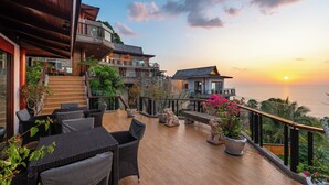 Een houten terras met buitenmeubilair, een trap en uitzicht op de oceaan bij zonsondergang.