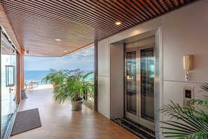 A modern hotel lobby with a wooden ceiling, a glass elevator, potted plants, and a view of the sea.