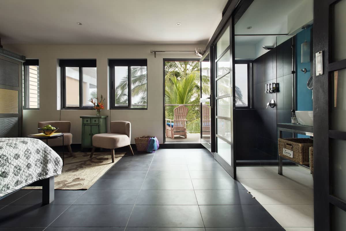 A modern bedroom with a bed, a small table with a vase, a chair, and a balcony with a view of palm trees.