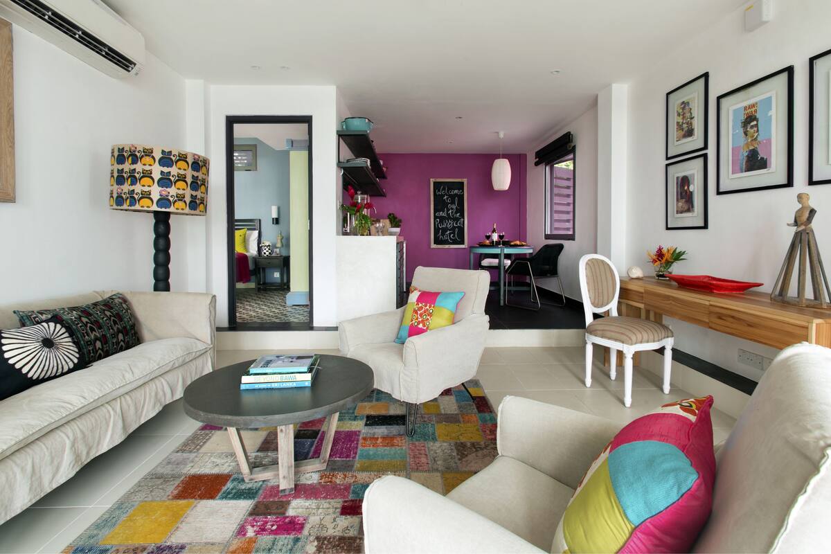 A modern living room with a sofa, armchairs, a coffee table, and a colorful rug. There is a dining area with a table and chairs, and a kitchen visible in the background.