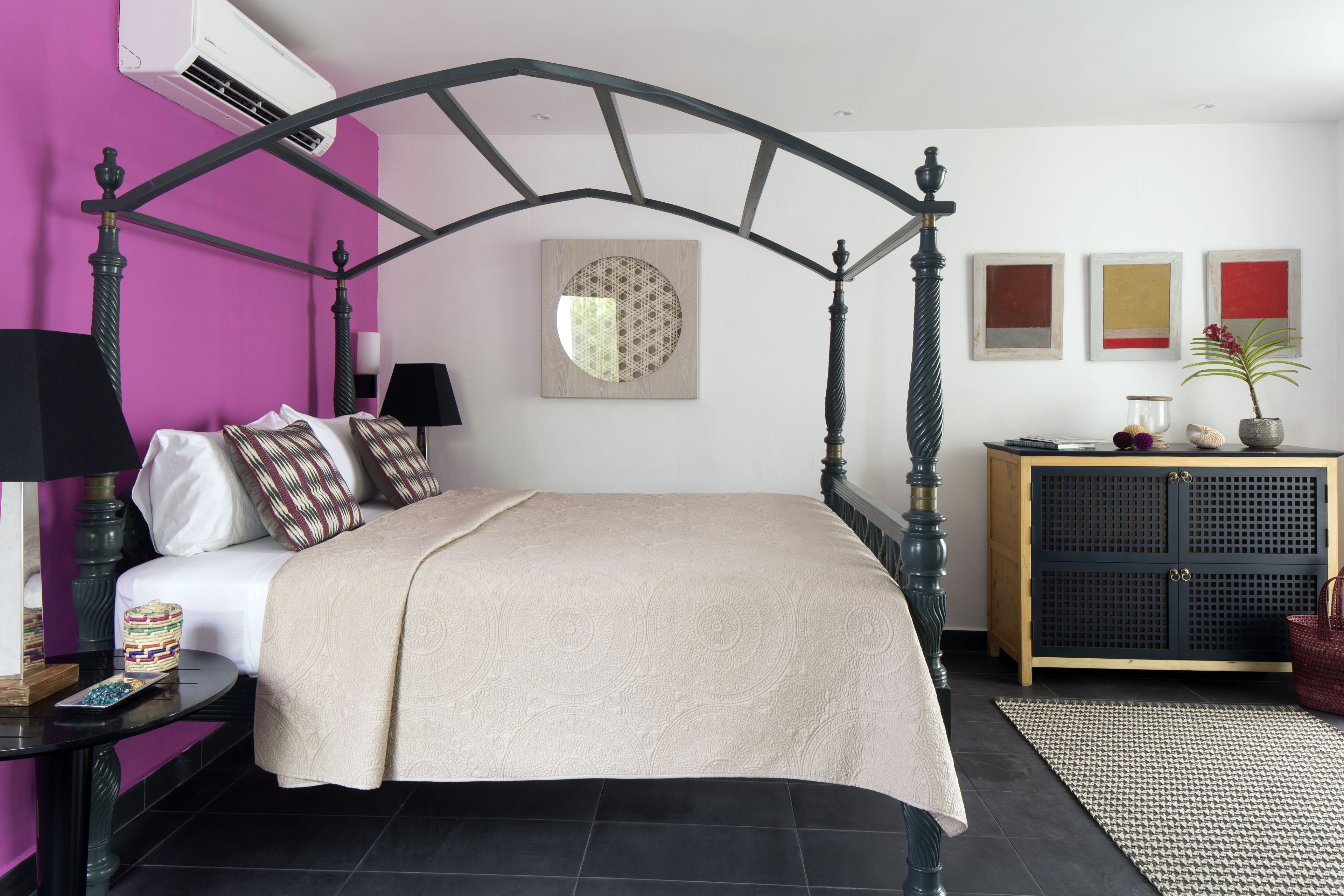 Una cama con dosel con cubrecolcha color beige, una mesita de noche de madera con lámpara, una mesita pequeña con un objeto decorativo y un aparador con una planta y objetos decorativos.