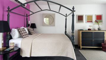 Una cama con dosel con cubrecolcha color beige, una mesita de noche de madera con lámpara, una mesita pequeña con un objeto decorativo y un aparador con una planta y objetos decorativos.