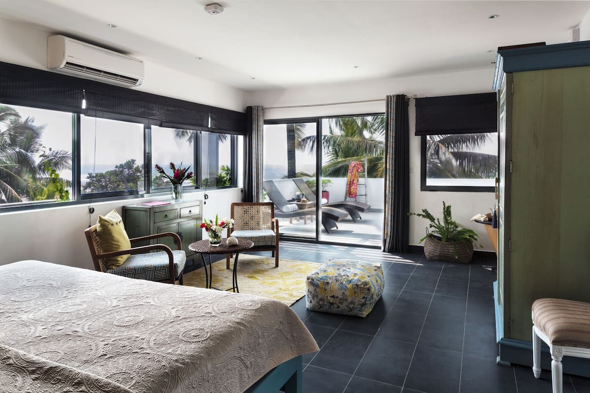 A spacious bedroom with a large bed, a sitting area with a chair and a small table, and a view of the outdoors through a sliding glass door.