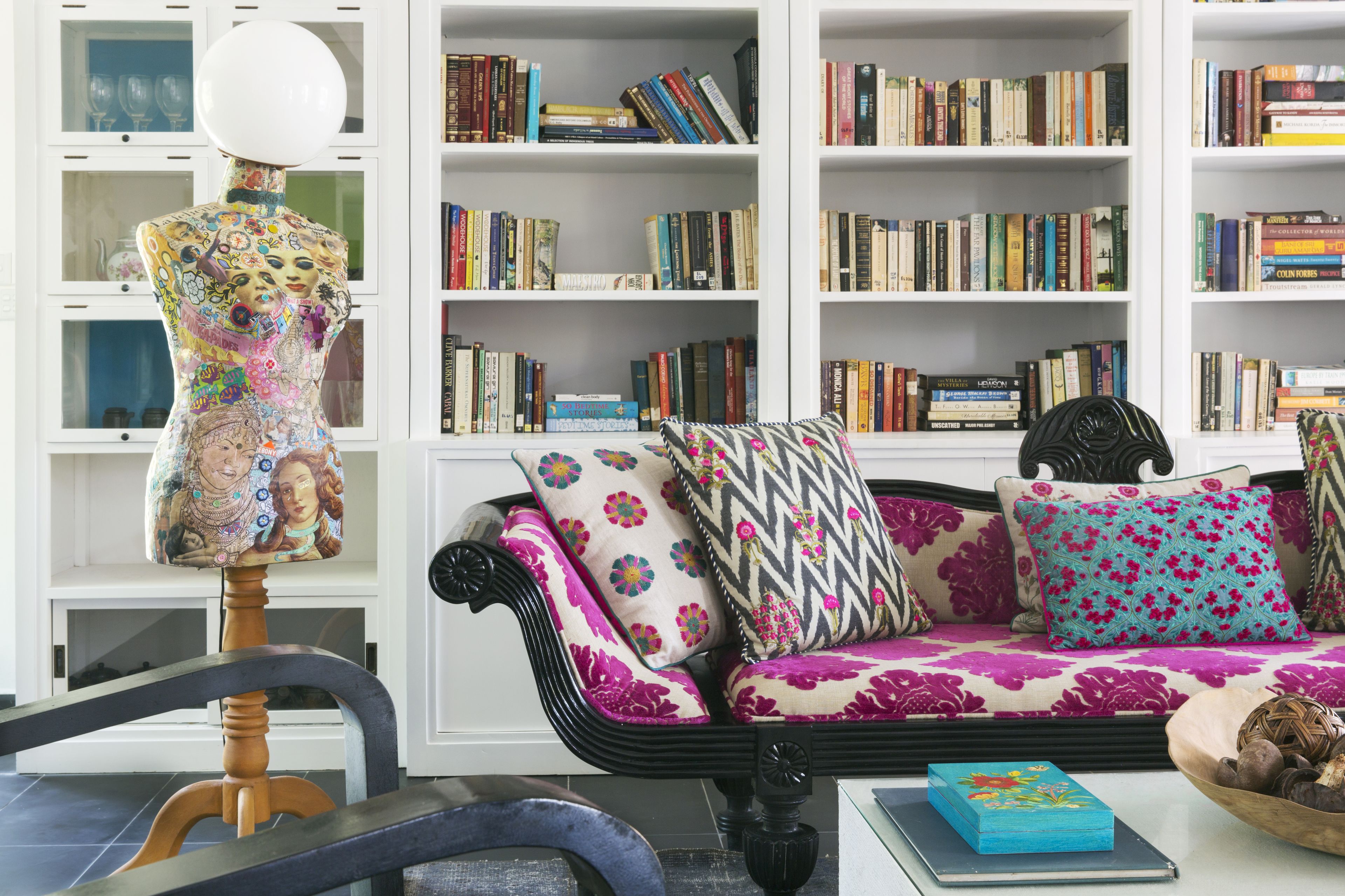 Una sala de estar con un sofá negro, cojines coloridos, una silla de madera y un maniquí. Se observan estanterías llenas de libros al fondo.