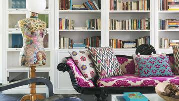 Una sala de estar con un sofá negro, cojines coloridos, una silla de madera y un maniquí. Se observan estanterías llenas de libros al fondo.