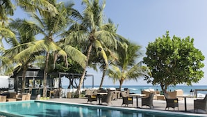 Un espace aménagé au bord de la piscine, avec des chaises longues et des tables, des palmiers et une vue sur l’océan.