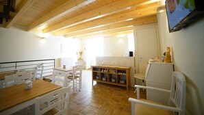 A dining area with wooden tables and chairs, a kitchen area with a counter and shelves, and a television mounted on the wall. - La Pergola ai Sassi (Matera)