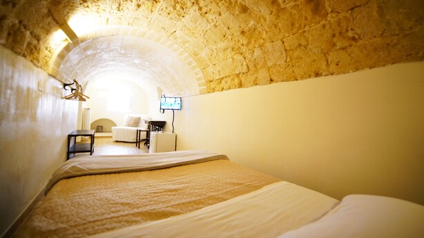 A room with a stone ceiling, a bed with a patterned bedspread, a small table, a chair, and a television on the wall. - La Pergola ai Sassi (Matera)