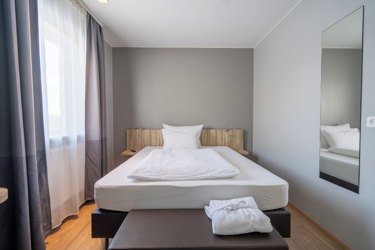 Un lit bien fait, avec du linge de lit blanc, une table de chevet sur laquelle est posée une serviette, et un grand miroir accroché au mur.