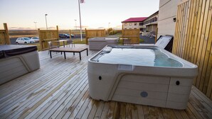 Un bain à remous sur une terrasse en bois, avec vue sur un immeuble et des voitures garées en arrière-plan.