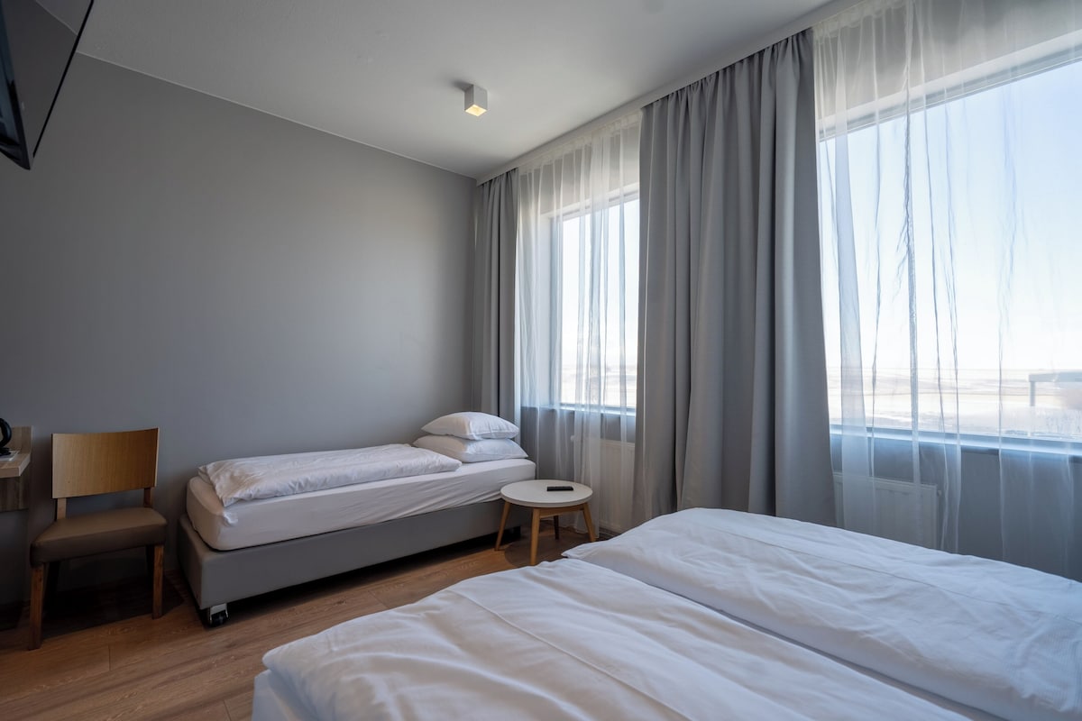 Une chambre d’hôtel avec un grand lit, un lit plus petit, une chaise, une table de chevet et une fenêtre avec des rideaux.