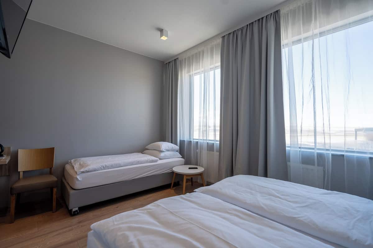 Une chambre d’hôtel avec un grand lit, un lit plus petit, une chaise, une table de chevet et une fenêtre avec des rideaux.