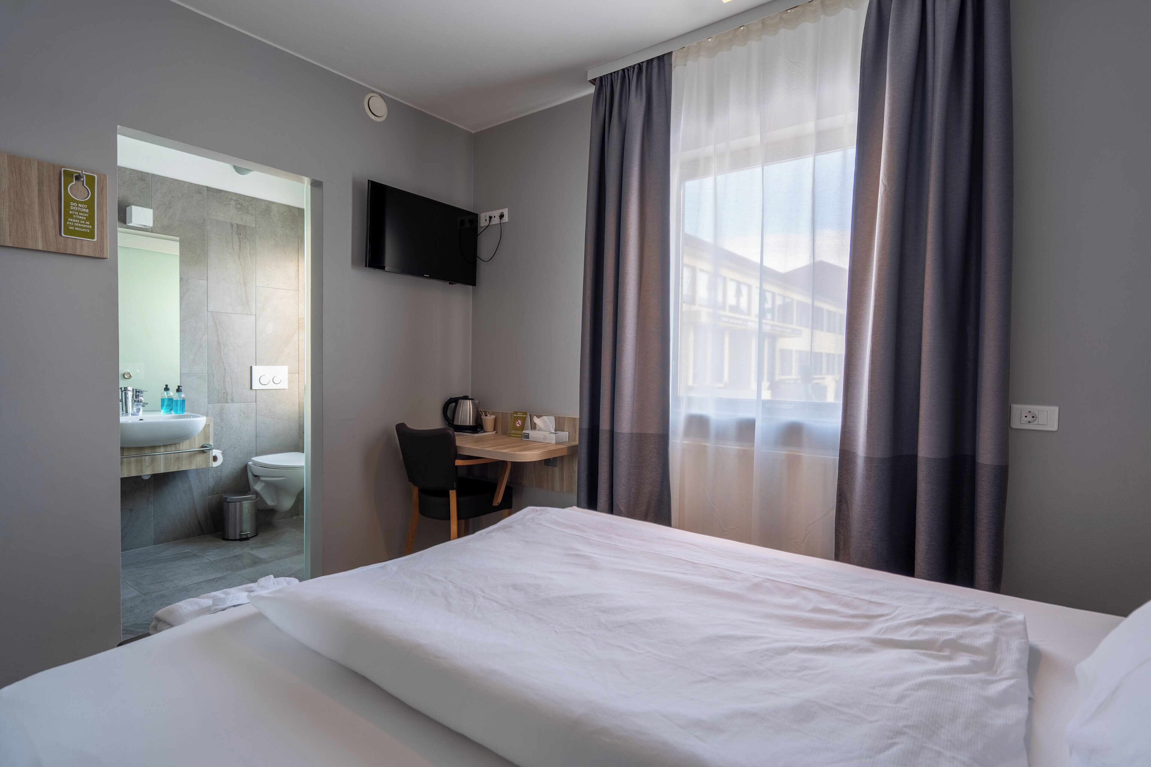 A hotel room with a bed, a desk, a chair, a TV, a bathroom with a sink and toilet, and a window with curtains.