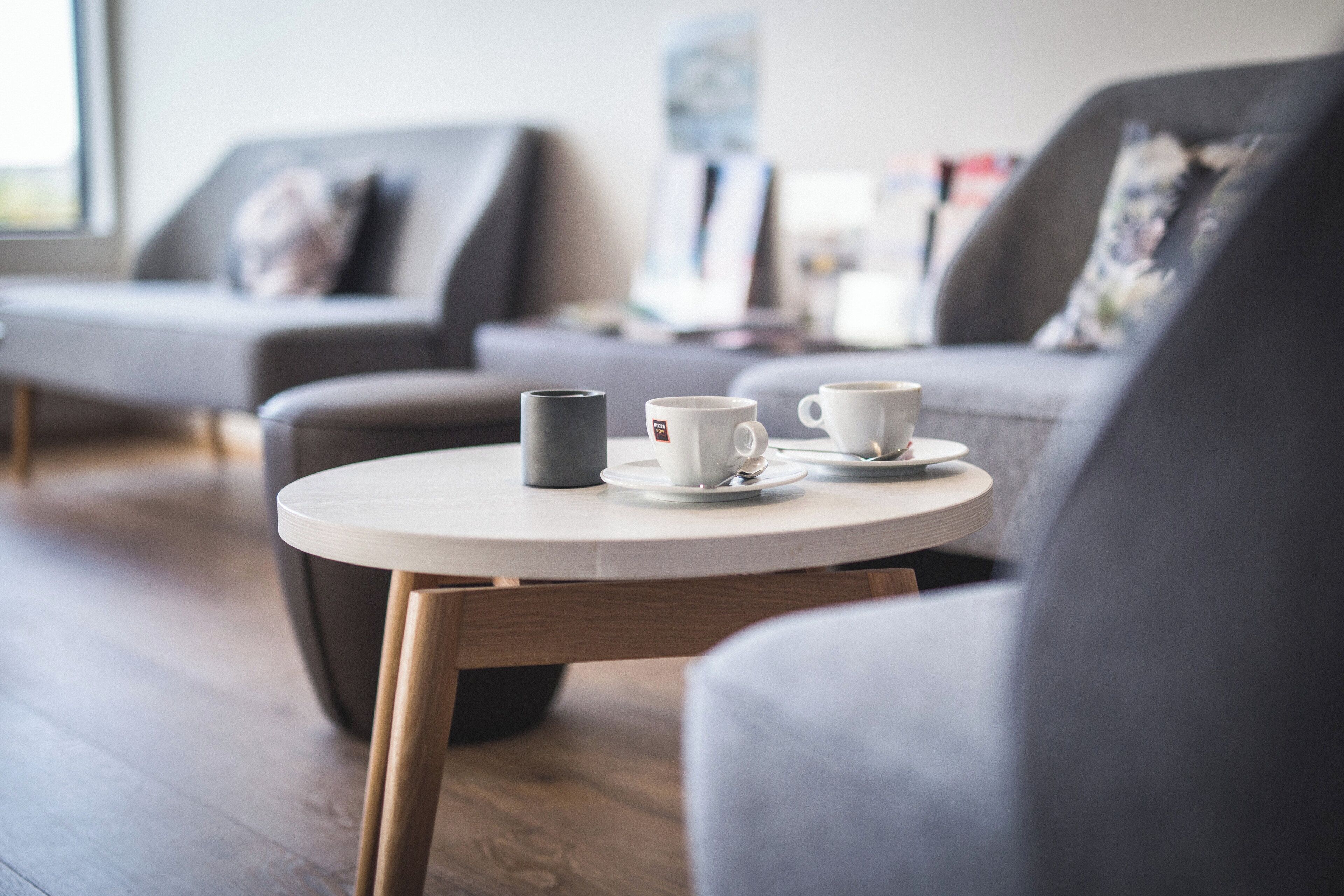A round table with two white cups and a grey mug, a grey sofa, and a wooden floor.