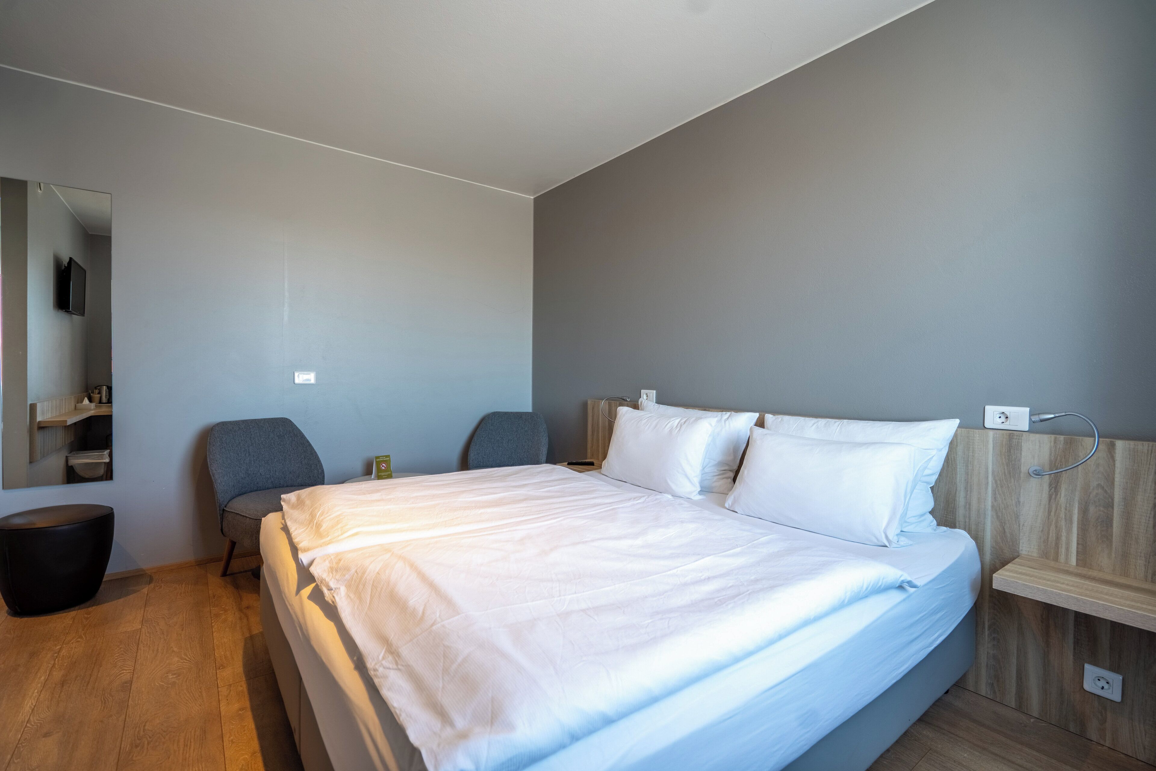 A modern hotel room with a large bed, a wooden floor, a gray wall, a small seating area, and a mirror.