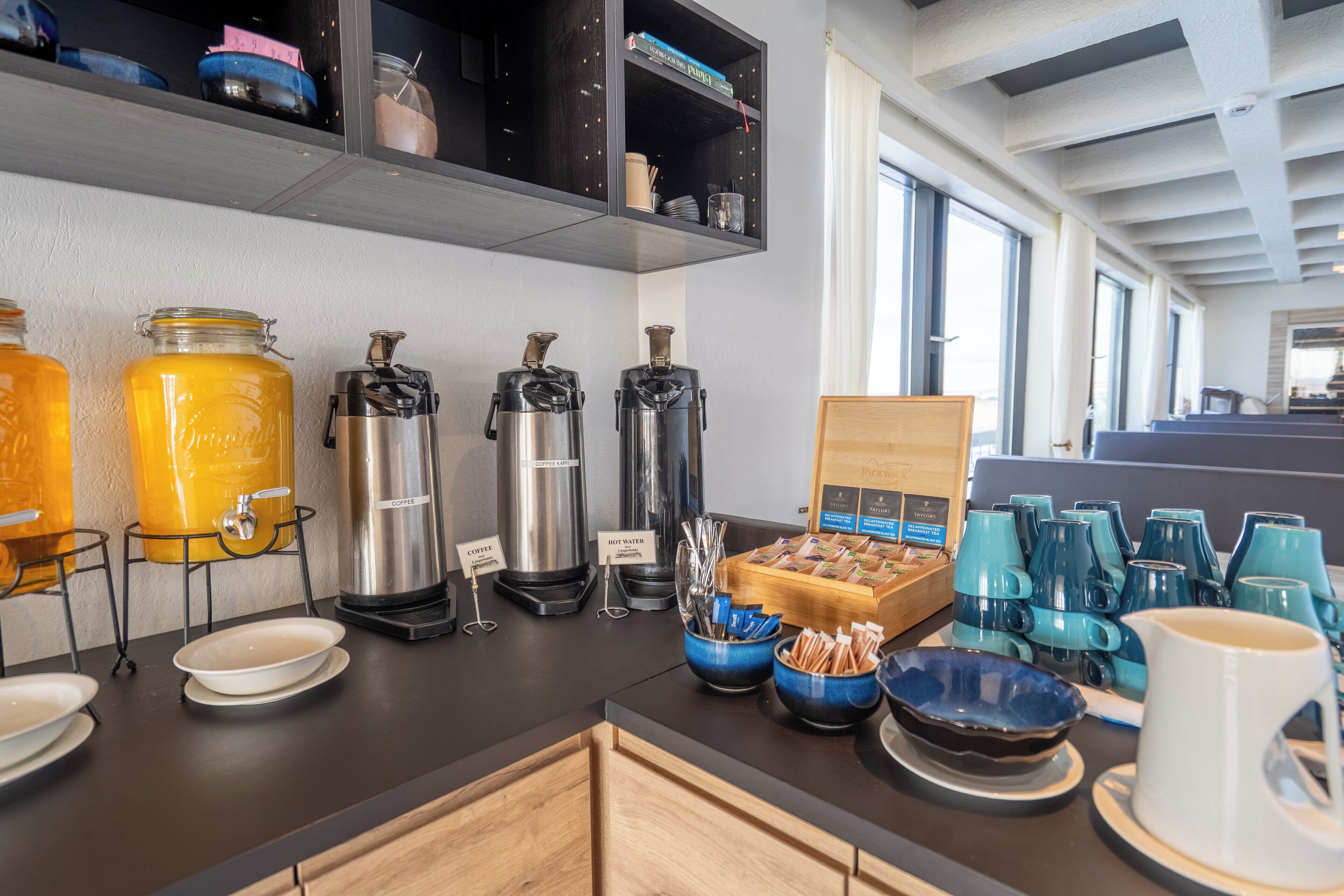 A breakfast bar with coffee dispensers, hot water station, and a variety of mugs and bowls.