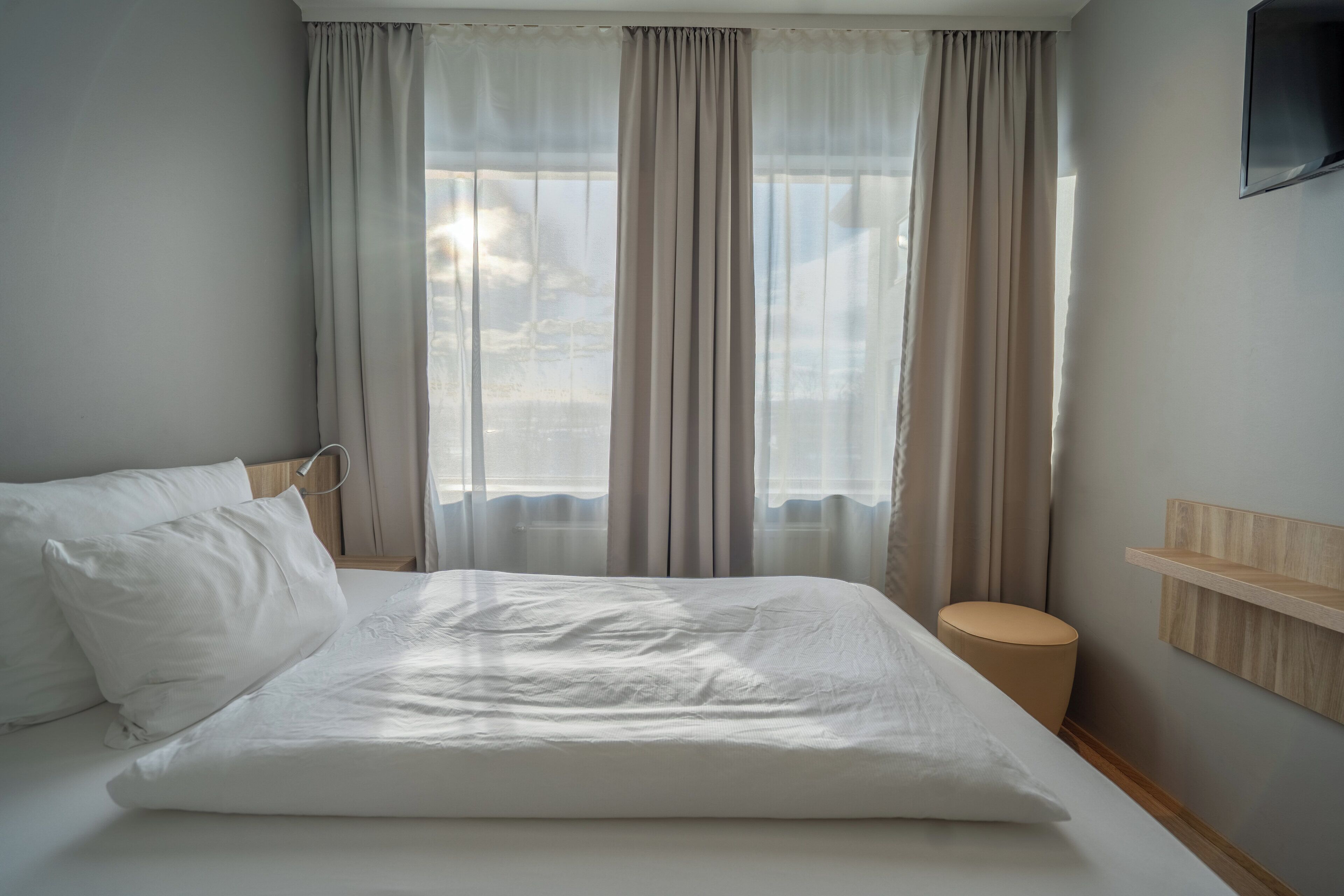 Una cama bien hecha con sábanas blancas, una mesita de noche de madera, un taburete pequeño y redondo, y un televisor montado en la pared.