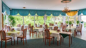 A spacious dining area with multiple tables and chairs, large windows offering a view of greenery, and a decorative chandelier. - Parkhotel Neustadt (Neustadt)