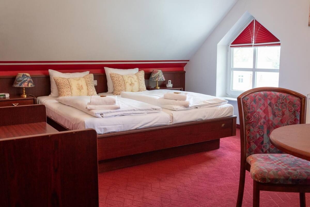 A hotel room with a large bed, two bedside tables with lamps, a chair, and a window with a red valance.