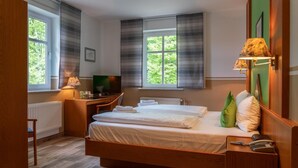 A hotel room with a wooden bed, a desk with a lamp, a television, and a window with blinds. - Parkhotel Neustadt (Neustadt)