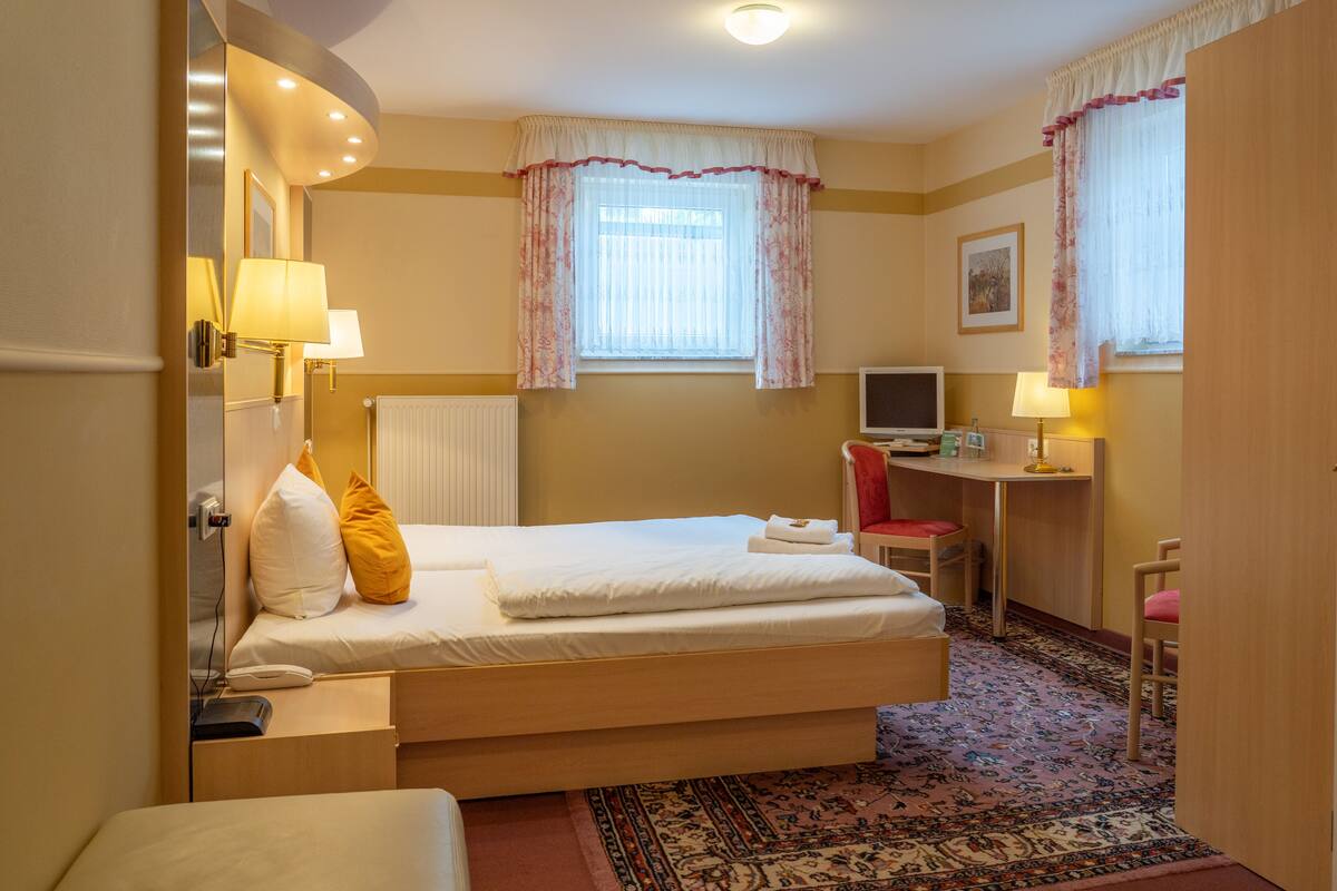 A hotel room with a bed, bedside table, lamp, desk with chair, and a television.