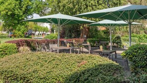 An outdoor patio area with tables and chairs under green umbrellas, surrounded by lush greenery and trees. - Parkhotel Neustadt (Neustadt)